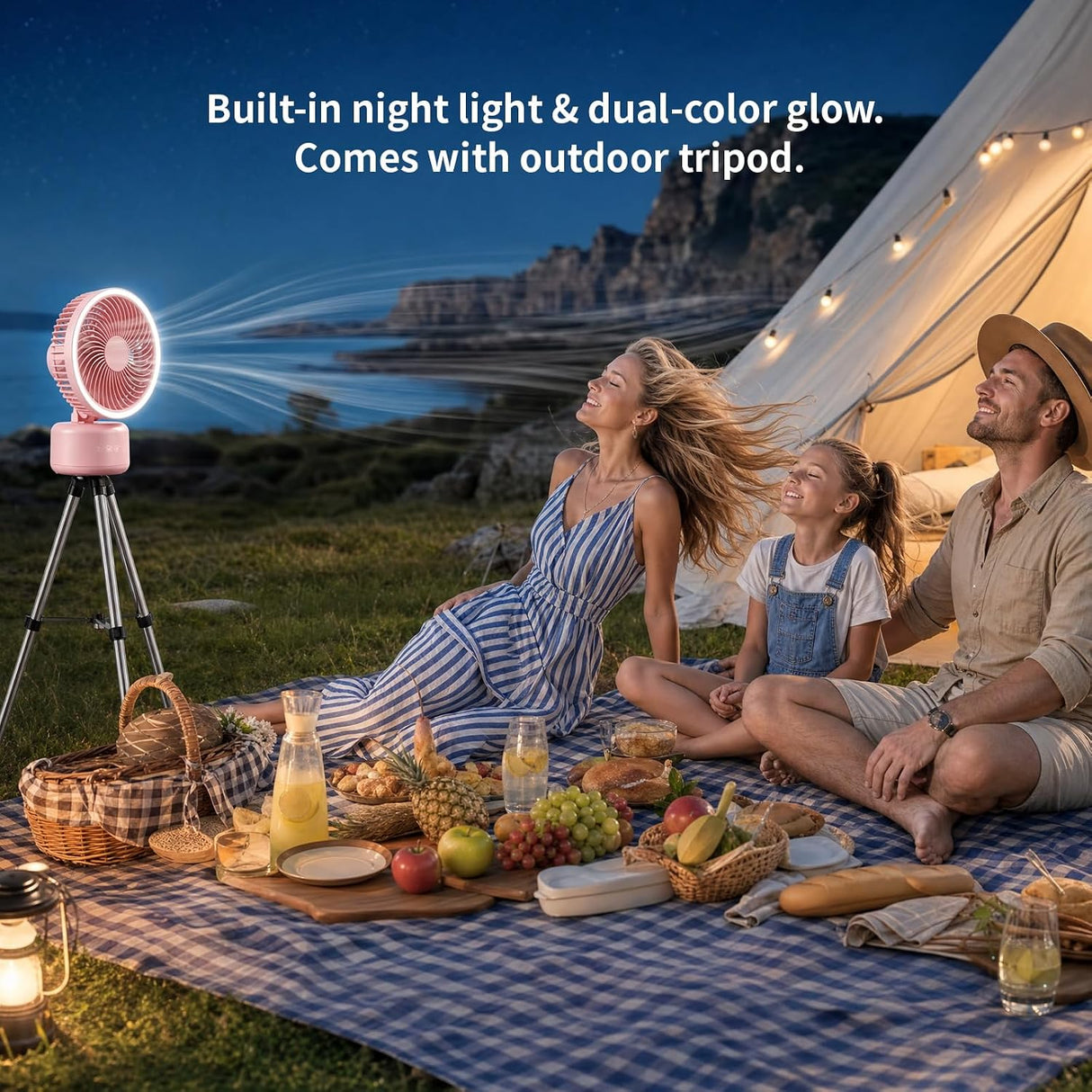 A family picnic outdoors at night beside a glowing tent, with a pink portable fan on a tripod casting light beams toward them. Text at the top reads: 'Built-in night light & dual-color glow.