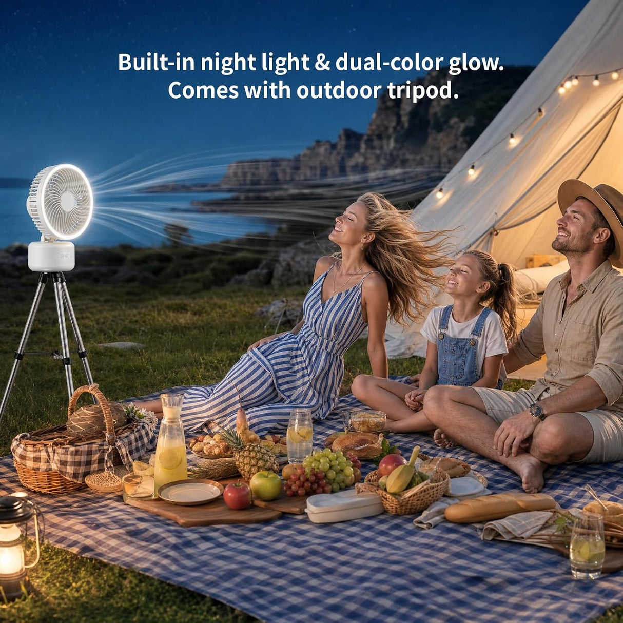 A family picnic outdoors at night under a string-tent canopy, with a portable white projector fan on a tripod casting a bright light beam across the sky. Text at the top reads, 'Built-in night light & dual-color glow.