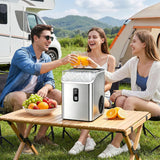 A stainless steel portable ice maker on a folding table outdoors, with three people toasting with glasses of orange juice in a camping setting with a tent, RV, and mountains in the background.