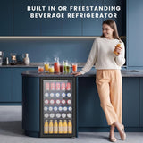 A woman stands beside a built-in beverage refrigerator in a modern kitchen setting. The refrigerator is dark blue and glass-front, stocked with bottled and canned drinks, with several drinks and pitchers on the countertop.
